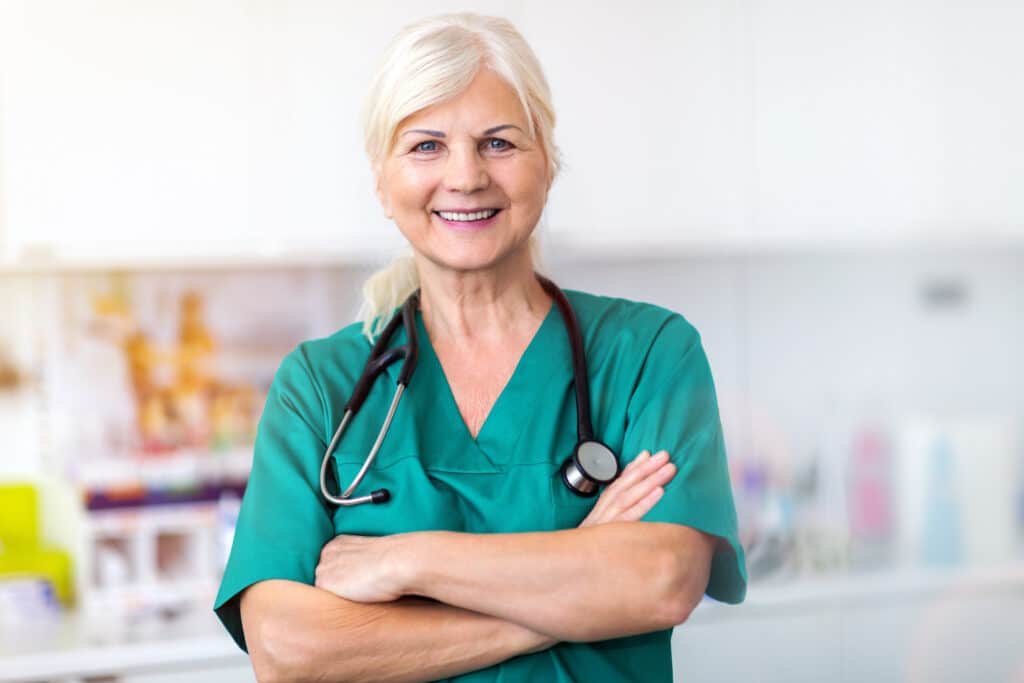 Eine blonde, ältere Ärztin in grünem OP Outfit lacht frontal in die Kamera.